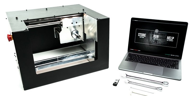 A "Ghost Gunner 3" CNC machine, sits next to a open laptop displaying Defence Distributed's website.